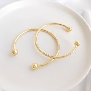 Op de afbeelding: Twee gouden armbandjes met ronde bolletjes aan de uiteinden.