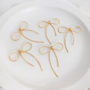 May include: Five gold-colored wire bow charms on a white background. The bows are made of thin wire and have a simple, elegant design.