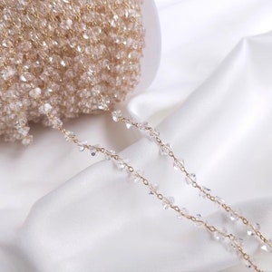 May include: A gold chain with clear crystal beads. The chain is coiled up on a white surface.