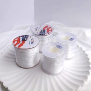 Peut inclure: Trois bobines de fil élastique blanc, chacune avec un récipient en plastique transparent et une étiquette indiquant "USA" avec un motif de drapeau américain.