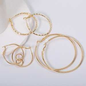May include: A collection of gold-colored hoop earrings in various sizes and designs. The earrings include smooth and textured hoops, with different clasp styles. The jewelry is displayed on a white surface.