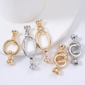 Könnte beinhalten: Eine Sammlung gold- und silberfarbener Schmuckverschlüsse in verschiedenen Designs. Die Verschlüsse umfassen runde, ovale und verschlungene Formen mit glänzendem, metallischem Finish. Die Verschlüsse werden auf einer weißen Oberfläche präsentiert.