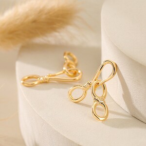 May include: Two gold-colored lobster claw clasps. The clasps have a rounded, elongated shape with a spring-loaded lever and a small ring at the end. The clasps are made of a shiny, reflective metal and are designed for jewelry making or crafts.