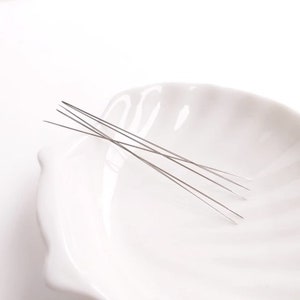 May include: A set of five thin, silver-colored needles in a white ceramic bowl.