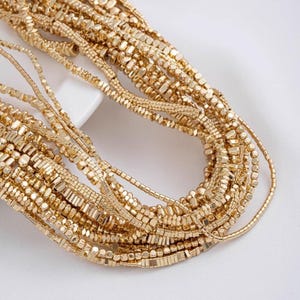 May include: An assortment of gold-colored necklaces with various bead shapes and sizes. The necklaces are arranged in a layered, cascading style, creating a rich, textured appearance. The beads include small squares, rectangles, and round shapes.