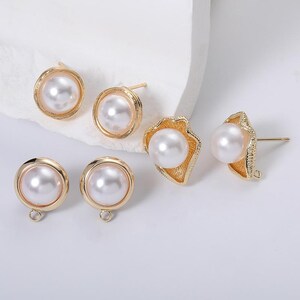 May include: A collection of gold-toned earrings featuring faux pearls. The earrings include classic round stud designs and a unique shell-shaped design. The pearls are white and the metal has a shiny finish. Some earrings have loops for attaching charms.