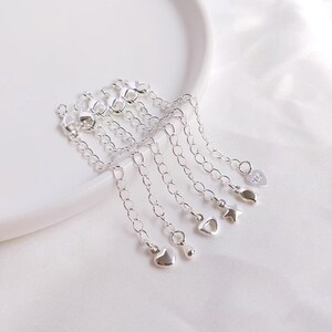 Könnte beinhalten: Ein Set aus sechs silbernen Kettenverlängerungen mit verschiedenen Anhängern, darunter Herzen, Sterne und eine Träne. Die Verlängerungen sind alle etwa 5 cm lang und perfekt, um Halsketten oder Armbändern eine persönliche Note zu verleihen.