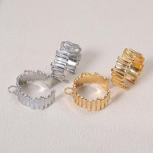 Puede incluir: Cuatro anillos abiertos en tonos plateados y dorados. Cada anillo presenta un diseño texturizado de barras verticales. Un anillo tiene un pequeño lazo adjunto. Los anillos se muestran sobre una superficie de color claro.