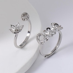 May include: Two silver-colored rings. One ring features a flower design, while the other has a heart and flower design. The rings are set against a white background, showcasing their intricate details and elegant style.