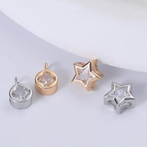 Op de afbeelding: Vier kleine, decoratieve bedels in zilver- en goudtinten. Twee bedels zijn stervormig en twee zijn klavervormig. Elke bedel heeft een heldere, facetgeslepen steen. De bedels worden op een wit oppervlak weergegeven.