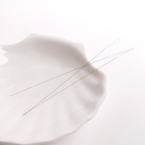 May include: Two thin, silver-colored threading needles are lying on a white ceramic dish. The dish is shaped like a seashell.