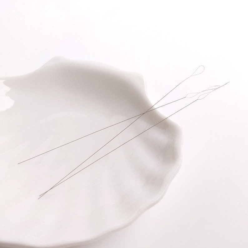 May include: Two thin, silver-colored threading needles are lying on a white ceramic dish. The dish is shaped like a seashell.