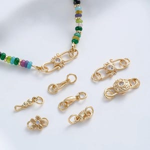 Puede incluir: Cierres y conectores de joyería dorados en varios diseños, incluyendo una flor, un cierre redondo y un cierre decorativo con una piedra transparente. También se ve un collar de cuentas con cuentas verdes, azules y moradas.