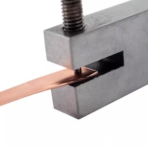 Puede incluir: Una herramienta de sujeción de metal con una tira de cobre insertada en la abertura. La abrazadera se utiliza para sujetar la tira en su lugar mientras se perfora un agujero.