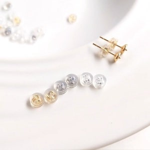 Puede incluir: Primer plano de un juego de respaldos para pendientes en diferentes colores, incluyendo oro, plata y transparente. Los respaldos están hechos de plástico y tienen una pequeña forma redonda.