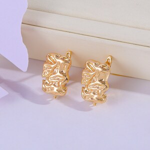 May include: A pair of gold-toned earrings with a textured, abstract design. The earrings are made of metal and have a hoop shape.