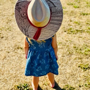 Pode incluir: Uma crian&ccedil;a usando um chap&eacute;u de cowboy claro com uma faixa vermelha e um vestido azul. O chap&eacute;u apresenta um desenho de uma vaca Highland e um celeiro. A crian&ccedil;a est&aacute; em um campo de grama seca.