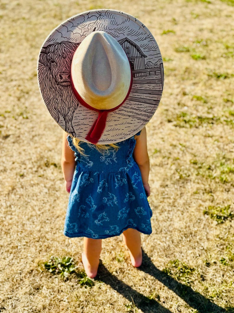 Pode incluir: Uma crian&ccedil;a usando um chap&eacute;u de cowboy claro com uma faixa vermelha e um vestido azul. O chap&eacute;u apresenta um desenho de uma vaca Highland e um celeiro. A crian&ccedil;a est&aacute; em um campo de grama seca.