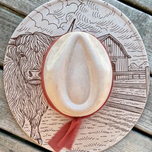 Pode incluir: Chap&eacute;u de cowboy bege com uma fita vermelha. O chap&eacute;u apresenta uma ilustra&ccedil;&atilde;o marrom de uma vaca das Terras Altas, um celeiro e uma paisagem rural. A aba do chap&eacute;u tem um design decorativo.