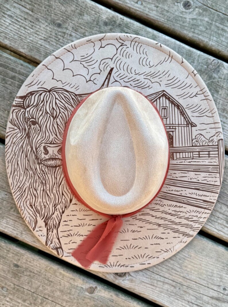 Pode incluir: Chap&eacute;u de cowboy bege com uma fita vermelha. O chap&eacute;u apresenta uma ilustra&ccedil;&atilde;o marrom de uma vaca das Terras Altas, um celeiro e uma paisagem rural. A aba do chap&eacute;u tem um design decorativo.