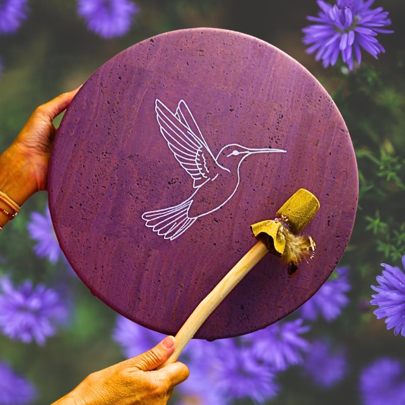 May include: A circular, purple drum featuring a white hummingbird outline. The drum is accompanied by a wooden drumstick with a yellow head and feathers. The background is a soft focus of purple flowers.