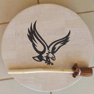 May include: A circular drum featuring a light beige surface, adorned with a black eagle illustration. A light brown drumstick with a brown fabric head lies across the drum. The drum is likely used for music or ceremonial purposes.