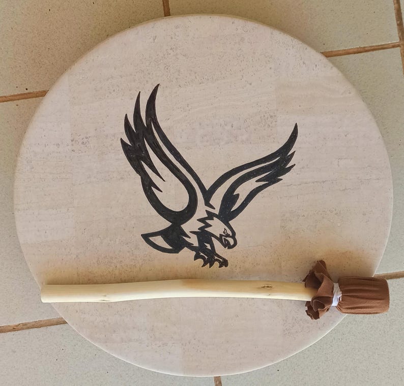 May include: A circular drum featuring a light beige surface, adorned with a black eagle illustration. A light brown drumstick with a brown fabric head lies across the drum. The drum is likely used for music or ceremonial purposes.