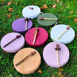 May include: A selection of handmade, circular drums in diverse hues, such as purple, pink, and green, placed on green grass. Each drum features a matching mallet with a brown head and a light-coloured handle.