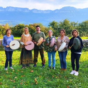 May include: Six people in a field, each holding a round drum with a beater. The drums are various colours, including white, pink, and green. Mountains are visible in the background under a cloudy sky. The people are standing in a grassy field.