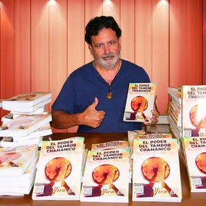 May include: A man in a blue shirt holds a book titled "EL PODER DEL TAMBOR CHAMANICO" with a circular graphic. Several copies of the book are stacked on a table. The background is a wooden wall with spotlights.