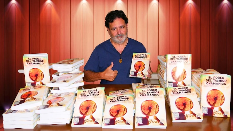 May include: A man in a blue shirt holds a book titled "EL PODER DEL TAMBOR CHAMANICO" with a circular graphic. Several copies of the book are stacked on a table. The background is a wooden wall with spotlights.