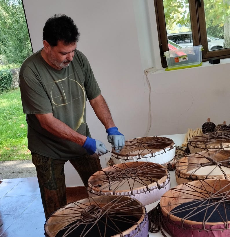 May include: A person crafting several handmade drums. The drums feature circular frames with stretched hides and detailed lacing. The drums are in various colours, including white, purple, and brown. The person is wearing a green shirt and blue gloves.