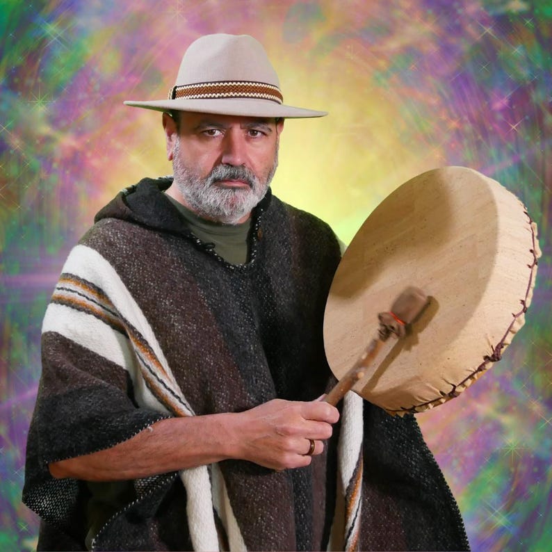 May include: A man wearing a beige hat with a brown band and a poncho with brown, white, and black stripes. He is holding a large, round drum with a wooden beater. The background has a colourful, blurred effect.