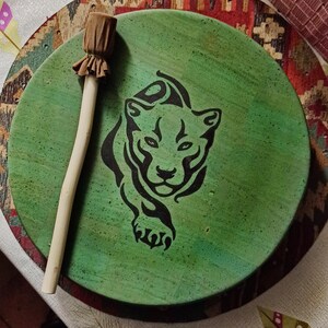 May include: A circular, green drum featuring a black panther design. It has a wooden handle and a brown fabric head. The drum is resting on a patterned surface.