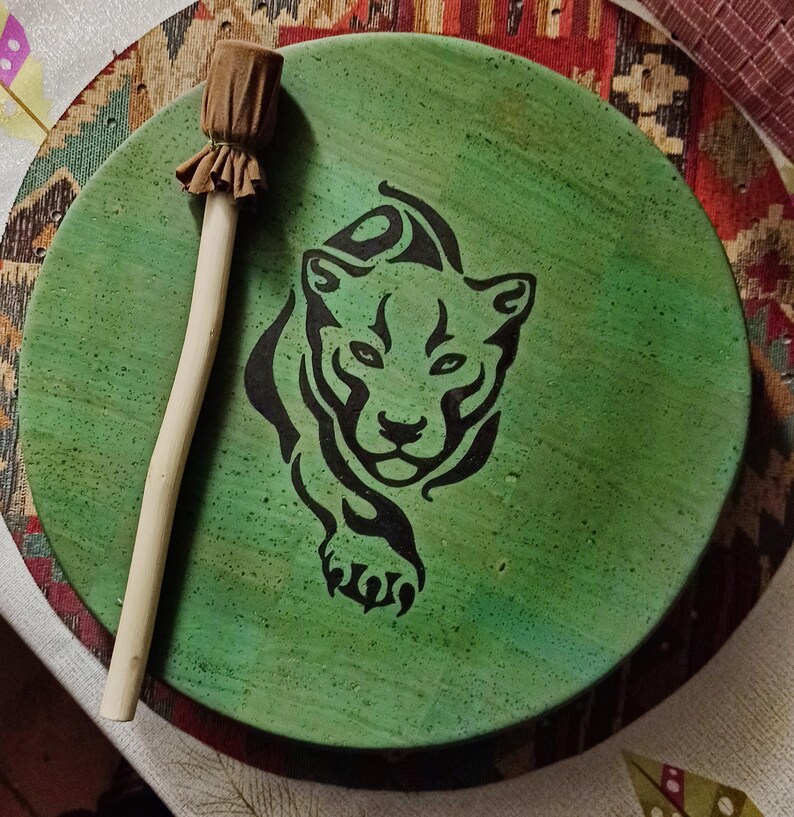 May include: A circular, green drum featuring a black panther design. It has a wooden handle and a brown fabric head. The drum is resting on a patterned surface.