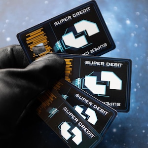 Könnte beinhalten: Eine Hand in einem schwarzen Handschuh hält mehrere schwarze und blaue Super Credit- und Super Debit-Karten. Jede Karte hat ein weißes Pfeildesign und die Wörter "Super Credit" oder "Super Debit" in Weiß. Der Hintergrund ist eine sternenklare, raumähnliche Szene.