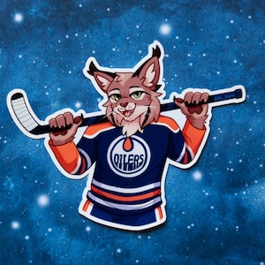 May include: A sticker featuring a cartoon lynx wearing an Edmonton Oilers hockey jersey and holding a hockey stick. The jersey is navy blue, orange, and white, with the Oilers logo on the chest. The background is a dark blue with white dots.