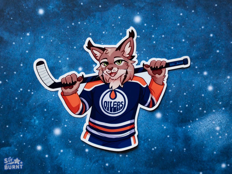 May include: A sticker featuring a cartoon lynx wearing an Edmonton Oilers hockey jersey and holding a hockey stick. The jersey is navy blue, orange, and white, with the Oilers logo on the chest. The background is a dark blue with white dots.