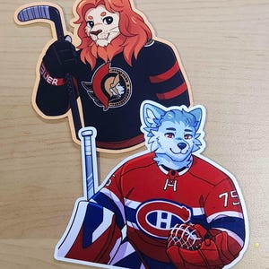 May include: Two stickers featuring anthropomorphic animals in hockey jerseys. One sticker shows a lion in a black jersey with red stripes, holding a hockey stick. The other sticker depicts a blue-furred wolf in a red jersey with a hockey glove.