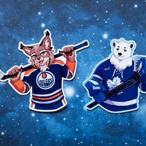 May include: Two die-cut stickers featuring anthropomorphic animals in hockey jerseys. One is a bobcat in an Oilers jersey, the other a polar bear in a Maple Leafs jersey, both holding hockey sticks. The background is a dark blue with white dots.