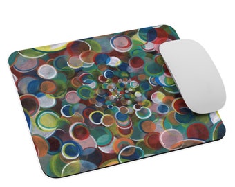 Può includere: Un tappetino per mouse rettangolare con un design astratto colorato di cerchi sovrapposti nei toni del rosso, blu, verde, giallo e bianco. Un mouse wireless bianco è posizionato a destra.