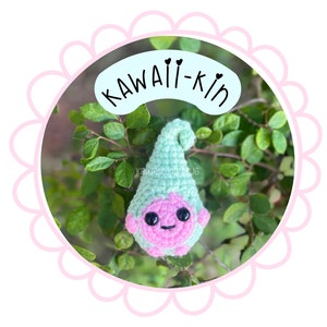 May include: A crocheted gnome with a pink body, green hat, and black eyes. The gnome is sitting on a green leafy branch. The text "KAWAII-KIN" is written in black on a pink circle.