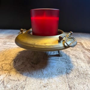 May include: A red candle in a brass candle holder. The candle holder has a circular design with three small legs and decorative handles. The candle is in a red glass votive.