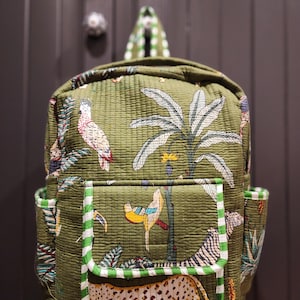 May include: A green quilted backpack with a jungle print featuring parrots, palm trees, and a cheetah. The backpack has a large front pocket with a green and white striped trim.