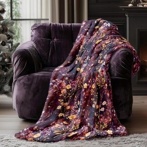May include: A plush throw blanket with a floral pattern in shades of pink, yellow, and orange on a dark blue background. The blanket is draped over a purple armchair in a living room setting.