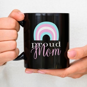 May include: Black coffee mug with a rainbow design in pink, blue, and white. The text "proud mom" is written in pink below the rainbow.