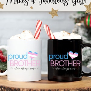 May include: Two mugs, one white and one black, with the words "proud BROTHER" and a heart in the colors of the transgender flag. The mugs are filled with marshmallows and candy canes. The text "love always wins" is also on the mugs.