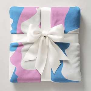 May include: A folded blanket with a white, blue, and pink abstract pattern, tied with a white satin ribbon and bow. The blanket is a square shape, and the bow is centered on the top.