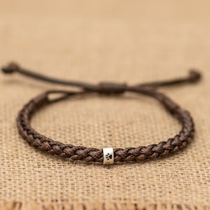 May include: A dark brown braided cord bracelet featuring a single cylindrical silver-toned bead. The bead is engraved with a small black paw print design. The bracelet includes adjustable cord ends for a secure fit around the wrist.