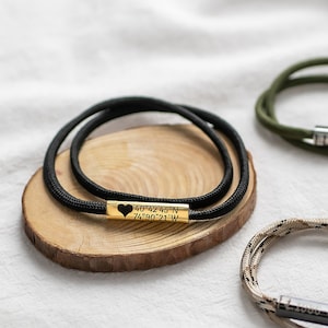 Puede incluir: Tres pulseras de paracord con cordones de diferentes colores y dijes de metal. Una pulsera tiene un dije de oro con coordenadas y un corazón, otra tiene un dije de plata con las palabras "Te amo", y la tercera tiene un dije de plata con una inscripción.